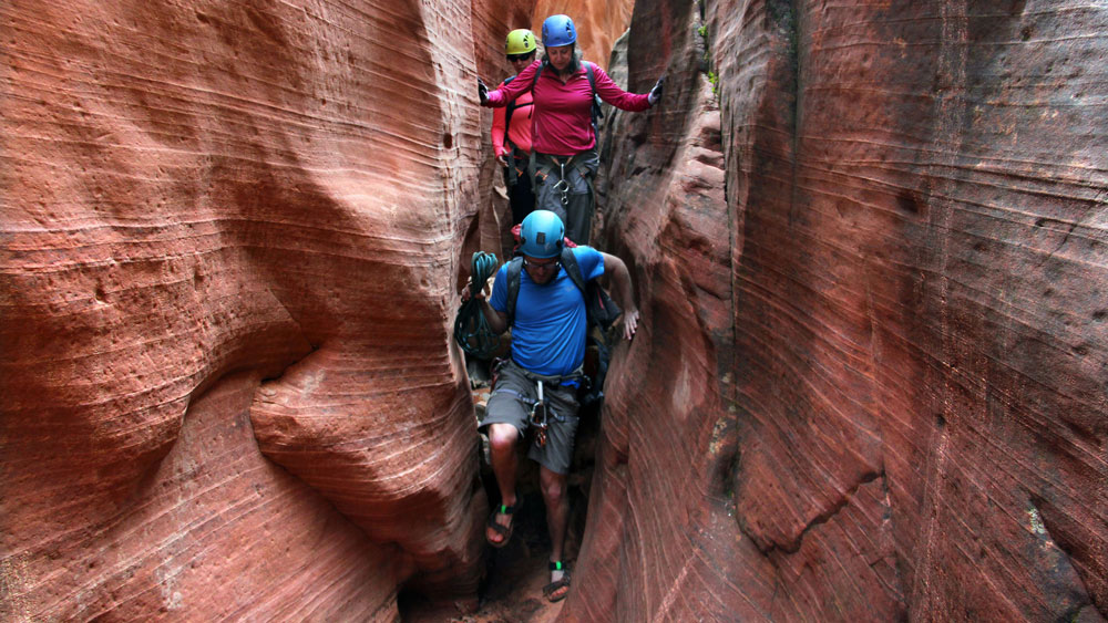 Three climbers navigating a narrow slot canyon with reddish rock formations.